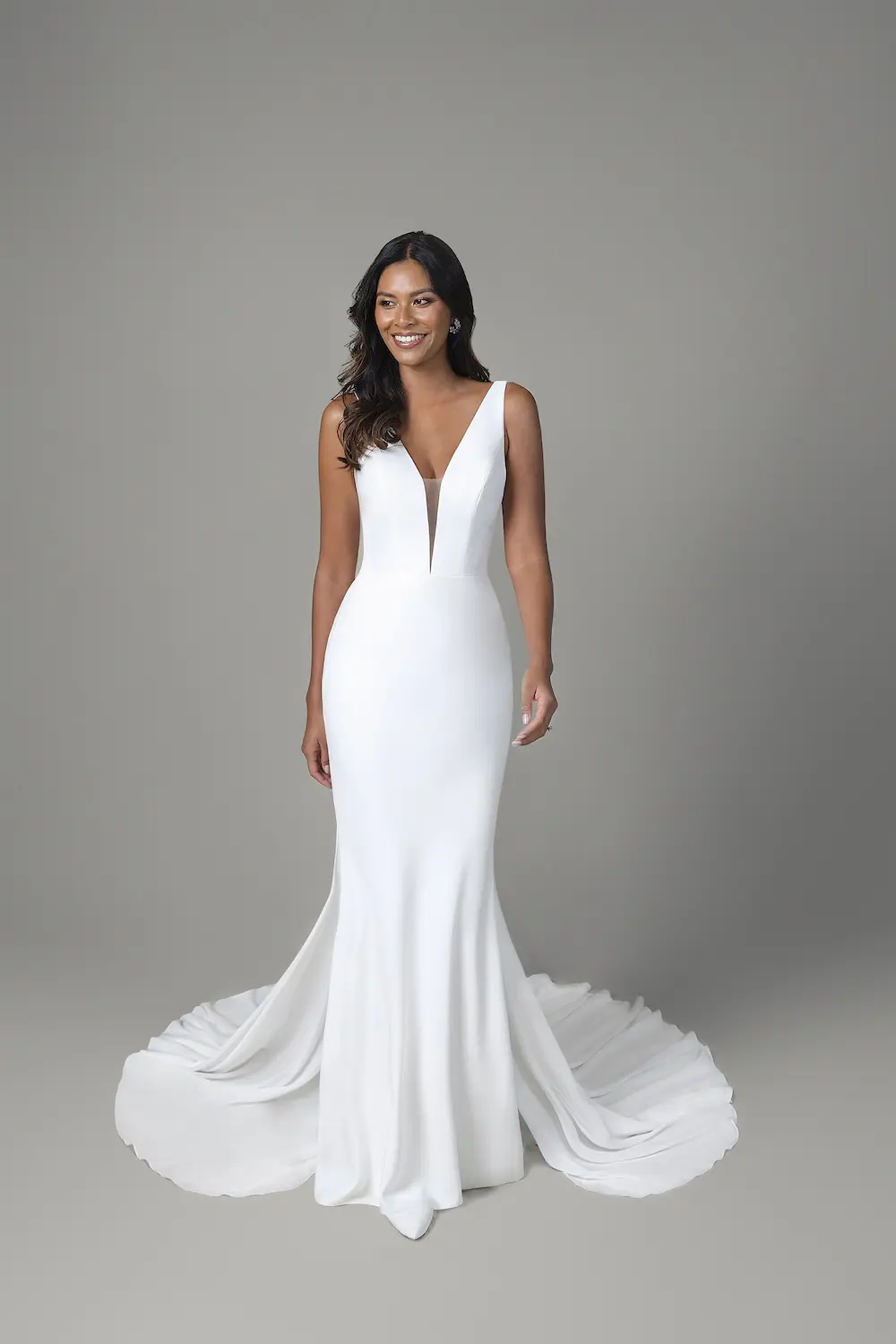 Rebecca, an ivory and latte fit-and-flare bridal gown by Sincerely Adorn, features a deep V-neckline with a sheer plunge inset. Crafted from smooth crepe, the gown is fitted through the bodice and hips, gracefully flaring into a dramatic chapel-length train. A woman with dark, wavy hair and a radiant smile models the dress, looking to the side.