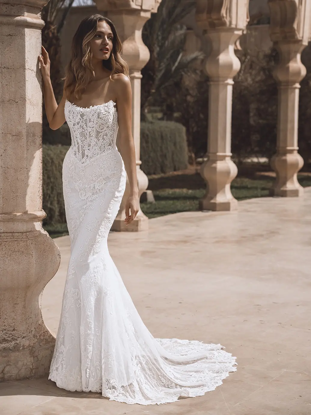 26-enzoani-pro-venice-d-fro-web Venice, an ivory and nude strapless mermaid wedding gown by Enzoani, features a vintage-inspired design crafted from intricate pearl-beaded floral and corded lace. The visible boning of the corset bodice sculpts the silhouette, which flares into a dramatic skirt and sweeping cathedral train. The bride stands elegantly beside a decorative stone column in a sunlit outdoor setting with arched colonnades and palm trees in the background, looking gently to the side.
