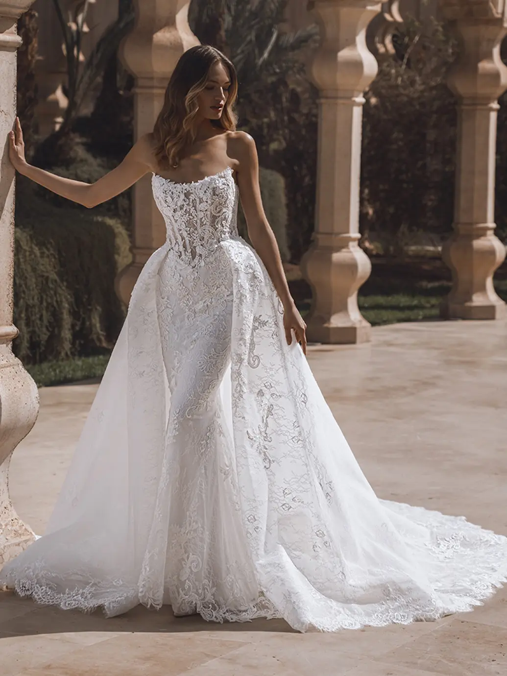 26-enzoani-pro-venice-front-web Venice, an ivory and nude Enzoani bridal gown, showcases a strapless, corset-style bodice intricately detailed with floral lace and delicate beading. The fit-and-flare silhouette is dramatically enhanced by a detachable A-line overskirt that cascades into a magnificent cathedral train. The gown also features buttons.
