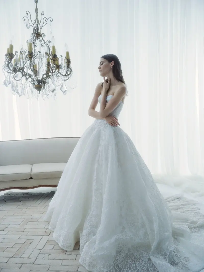 rivini-radiance-fw26-bridal-gown Radiance, an ivory diaphanous Basque waist ballgown crafted in delicate Chantilly corded lace for an ethereal effect, with a strapless corset bodice softly veiled in draped tulle and adorned with hand-appliquéd lace. The model, wearing the gown with a sweeping cathedral train, stands elegantly in a bright room with a chandelier, white sheer curtains, and a white sofa.