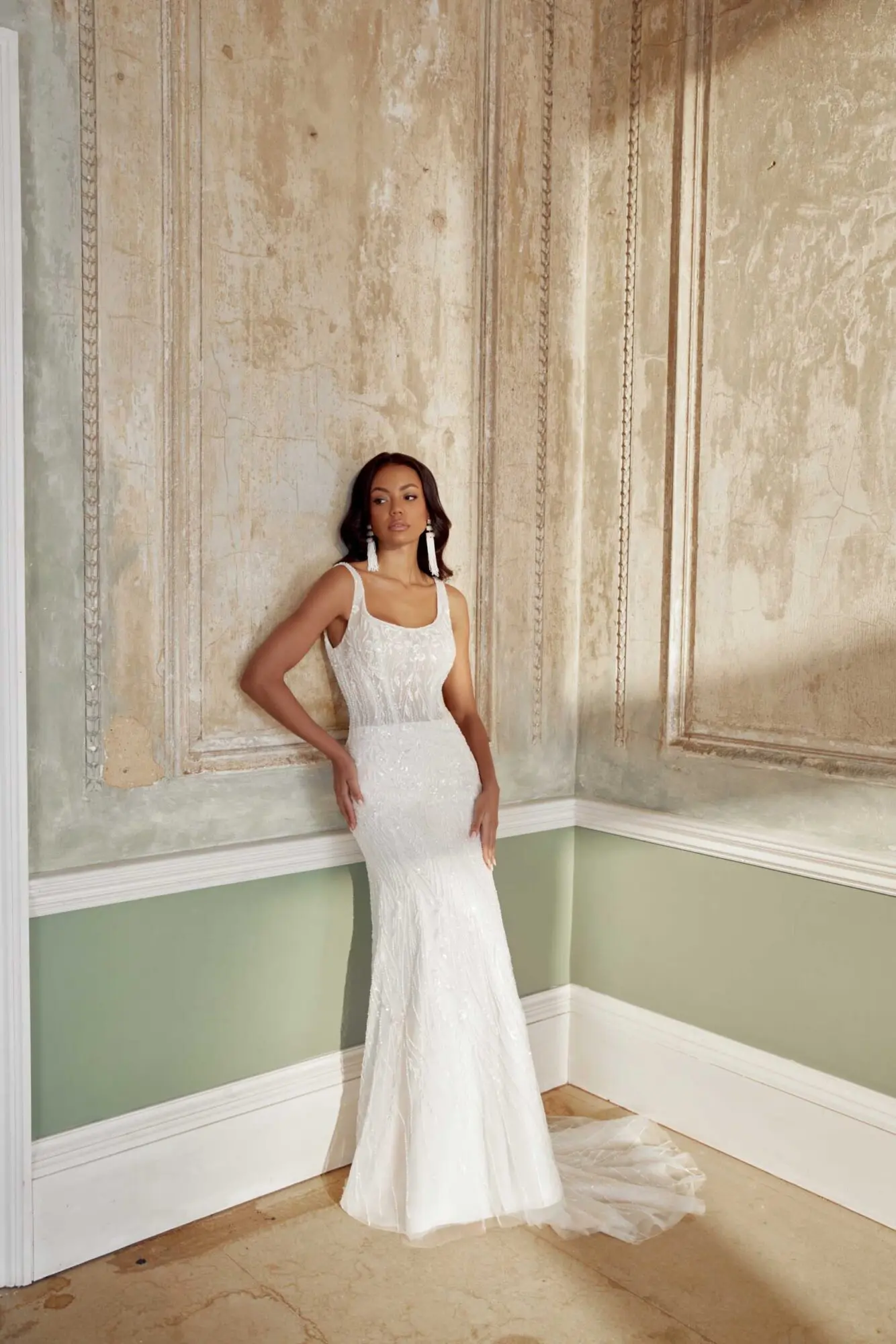Bridal Gown 44563, a Sand/Ivory/Latte fit-and-flare wedding gown with a demi sheer bodice and scoop neckline, is worn by a model leaning against an aged wall. The dress is richly embellished with lace, tulle, beading, embroidery, and glitter, flowing into a chapel train. The model, with dark hair, stands elegantly in a distressed room with cream and sage green walls, showcasing the gown's shimmering texture and silhouette.