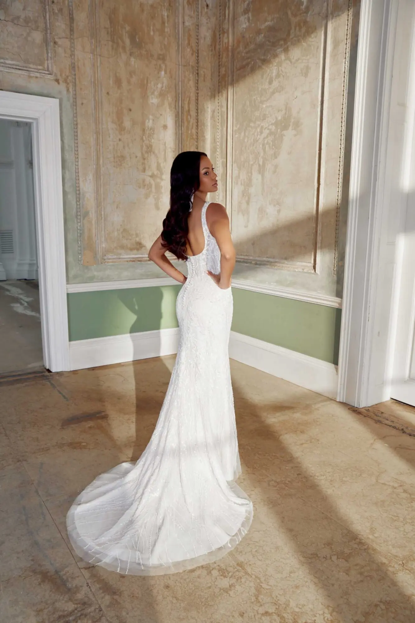 sincerity-bridal-style-88 A model in a shimmering ivory fit-and-flare wedding gown, posing with hands on hips, her back slightly turned and looking over her left shoulder. The gown features a demi-sheer scoop neckline bodice intricately embellished with glitter tulle, embroidered lace, and sparkling allover beadwork of seed beads, bugle beads, paillettes, and pearls. Covered buttons trail down the back to a chapel-length train. She stands in a sunlit room with distressed tan and green walls.