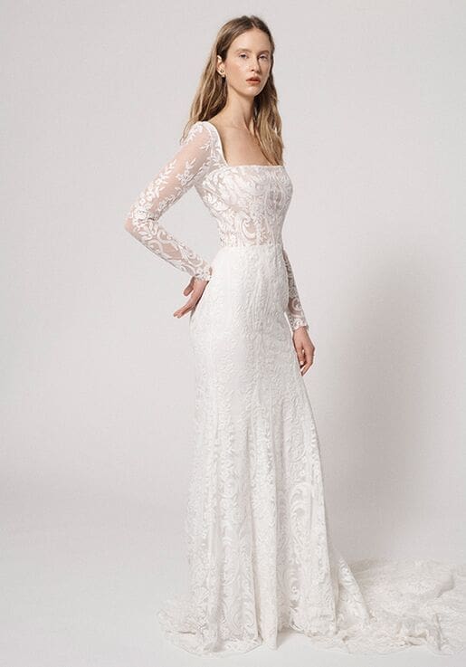 1616279415587-5eb03de6-34e9-4d6d-81e0-3b0fd7872356 Olivia, an ivory fit-and-flare wedding gown crafted entirely from intricate lace, features a demi-sheer bodice with a square neckline and long sleeves. The gown gently flares into a chapel-length train.