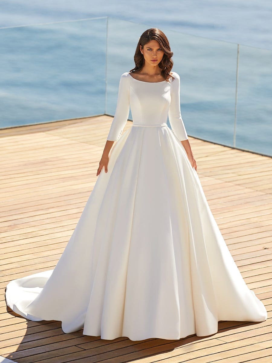 1627487910219-hope_b_22jzrd3cptoe3ebo Hope, an off-white A-line bridal gown by Pronovias, featuring a classic bateau neckline, modest 3/4-length sleeves with button details, and a delicate thin belt. The smooth satin fabric flows into a dramatic cathedral train.