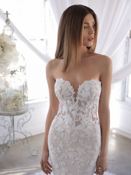 1631978650737-blue-22-pro-orrie-close-web-419-558-s-c1 Orrie, an ivory and sand/nude mermaid wedding gown with delicate, thick embroidered floral lace covering a sheer illusion bodice and a deep sweetheart neckline. The strapless dress is modeled by a woman with long brown hair, looking to the side, in a bright room with white drapes and a decorative floral arrangement.