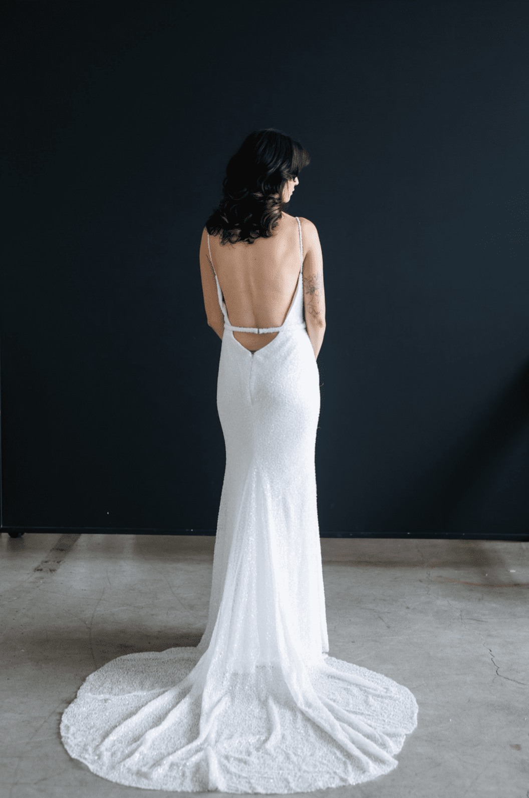 1668785610251-screen-shot-2022-11-18-at-10-32-50-am-2 Luna, an ivory fit-and-flare bridal gown by Hannah Hill, featuring shimmering beads and sequins, delicate beaded spaghetti straps, a sweetheart neckline, and an open, low back with a chapel-length train, shown from the back against a dark background.