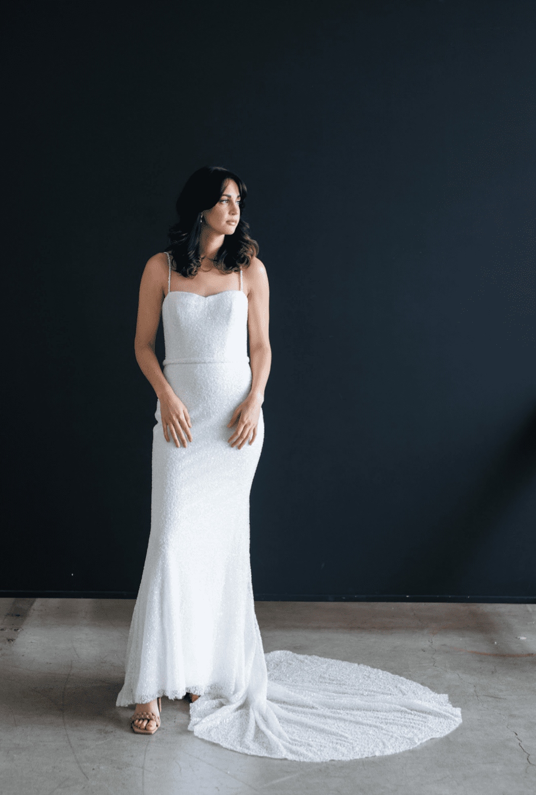 1668785610252-screen-shot-2022-11-18-at-10-33-13-am-1 Luna, a dazzling ivory fit-and-flare bridal gown covered entirely in shimmering beads and sequins, features a sweetheart neckline with delicate beaded spaghetti straps, an open low back, and a chapel-length train. The gown is modeled by a woman looking to her right against a dark background.