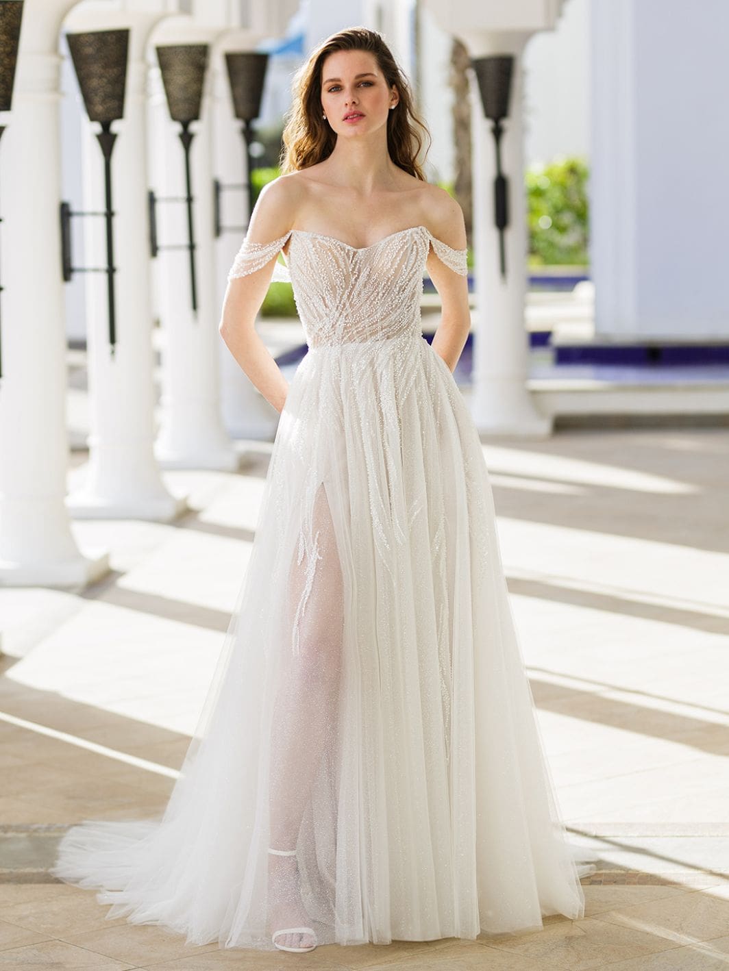 1686848059641-C62DAC6D-B6CD-437E-8DD4-BDAF8E9DF48F-2 Ruby, an ivory, blush, and nude A-line bridal gown, features a shimmering beaded lace bodice with a modified sweetheart neckline, illusion side cutouts, and delicate detachable off-the-shoulder beaded straps. The soft tulle skirt has a high slit revealing a glitter tulle layer and the model's leg, extending to a chapel train. The gown is worn by a woman posing in a sunlit outdoor corridor with white pillars.