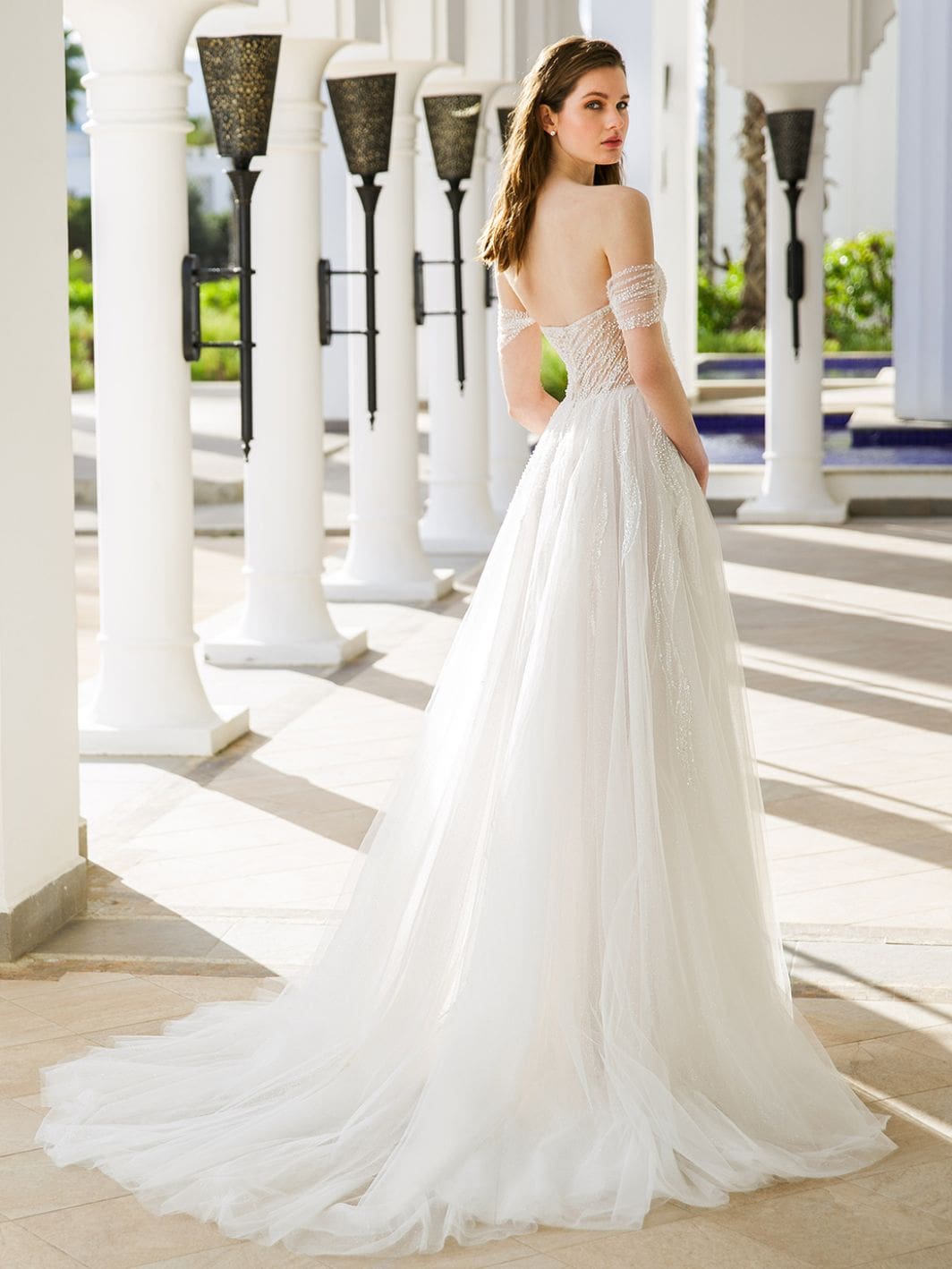 1686848059642-E06B6B8C-1786-4ACB-A5B2-8EB544B141FD-2 Ruby, an ivory, blush, and nude A-line bridal gown featuring a sculpted modified sweetheart neckline, illusion side cutouts, and ornate detachable off-the-shoulder beaded straps. The bodice showcases flowing beaded lace that extends into a voluminous tulle skirt, shimmering with glitter and a cascading lace overlay, forming a chapel-length train. The model, with her back slightly turned, gazes elegantly over her shoulder in an outdoor colonnade.
