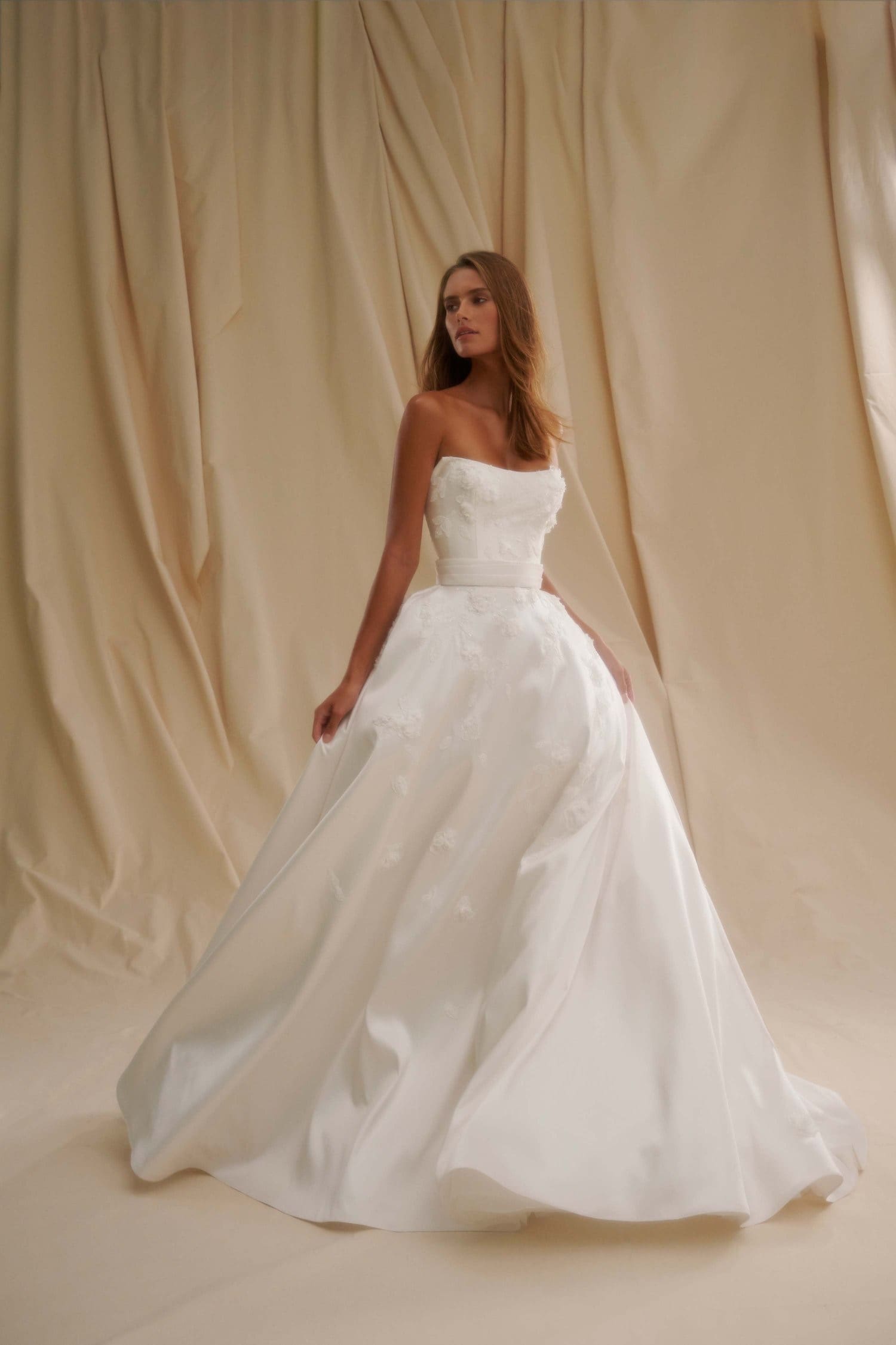1719681040875-image-9-2 A model in a Diamond White Senstudio A-line bridal gown, crafted from luxurious mikado fabric. The strapless dress features a cat-eye neckline, delicate 3D floral appliqués, and a sleek waist belt. She stands gracefully, looking to her left, against a softly draped cream background.