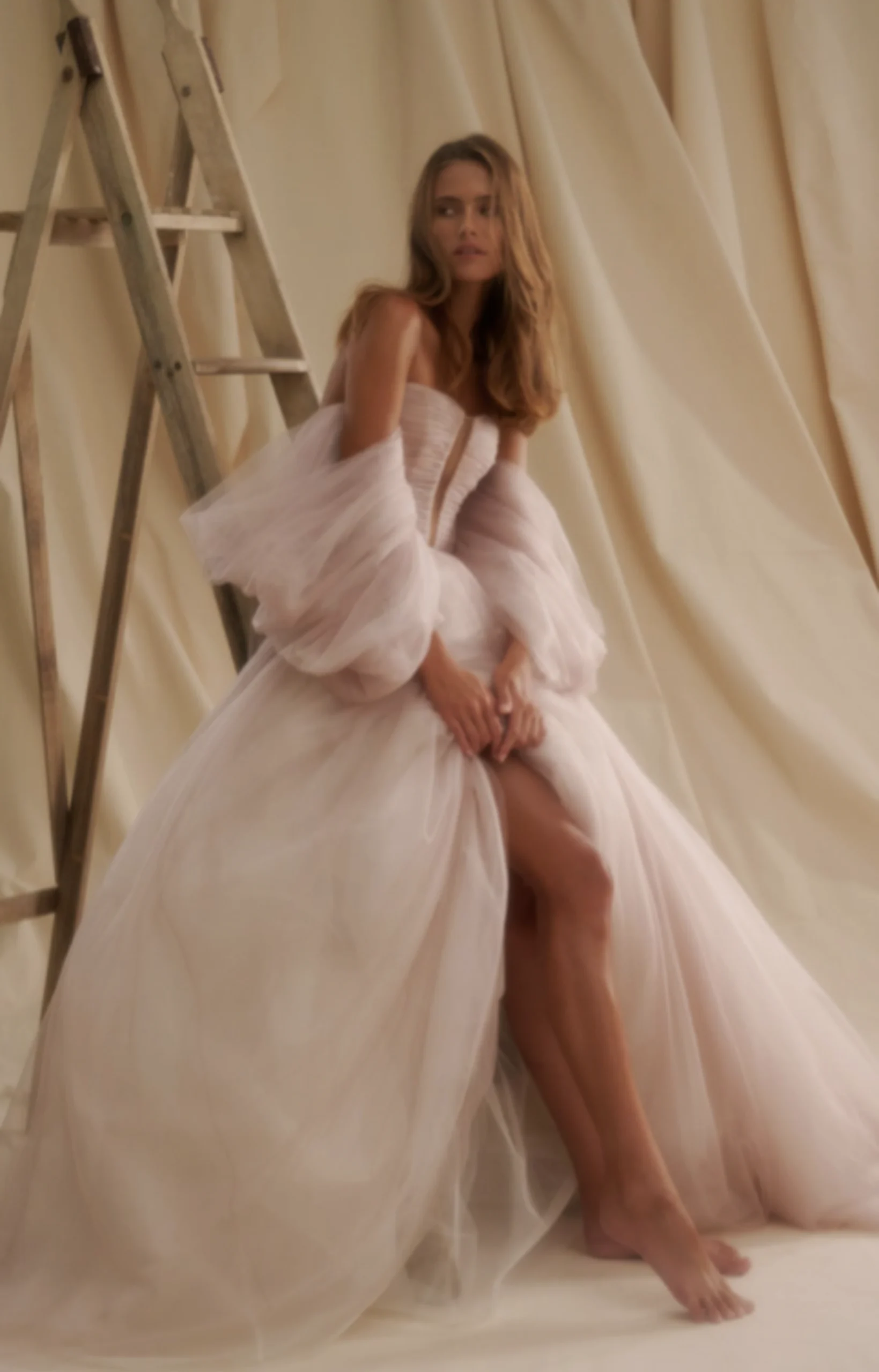 1720731980117-rv5h7hiytup0a3lypapf-2 Senstudio SE008, an ivory A-line bridal gown, is worn by a woman with long hair who is seated barefoot. The dress features a plunging strapless pleated bodice, delicate billowy tulle sleeves, and a flowing skirt with a high slit revealing her leg. A soft, draped fabric background and a wooden ladder complete the warm-toned scene.