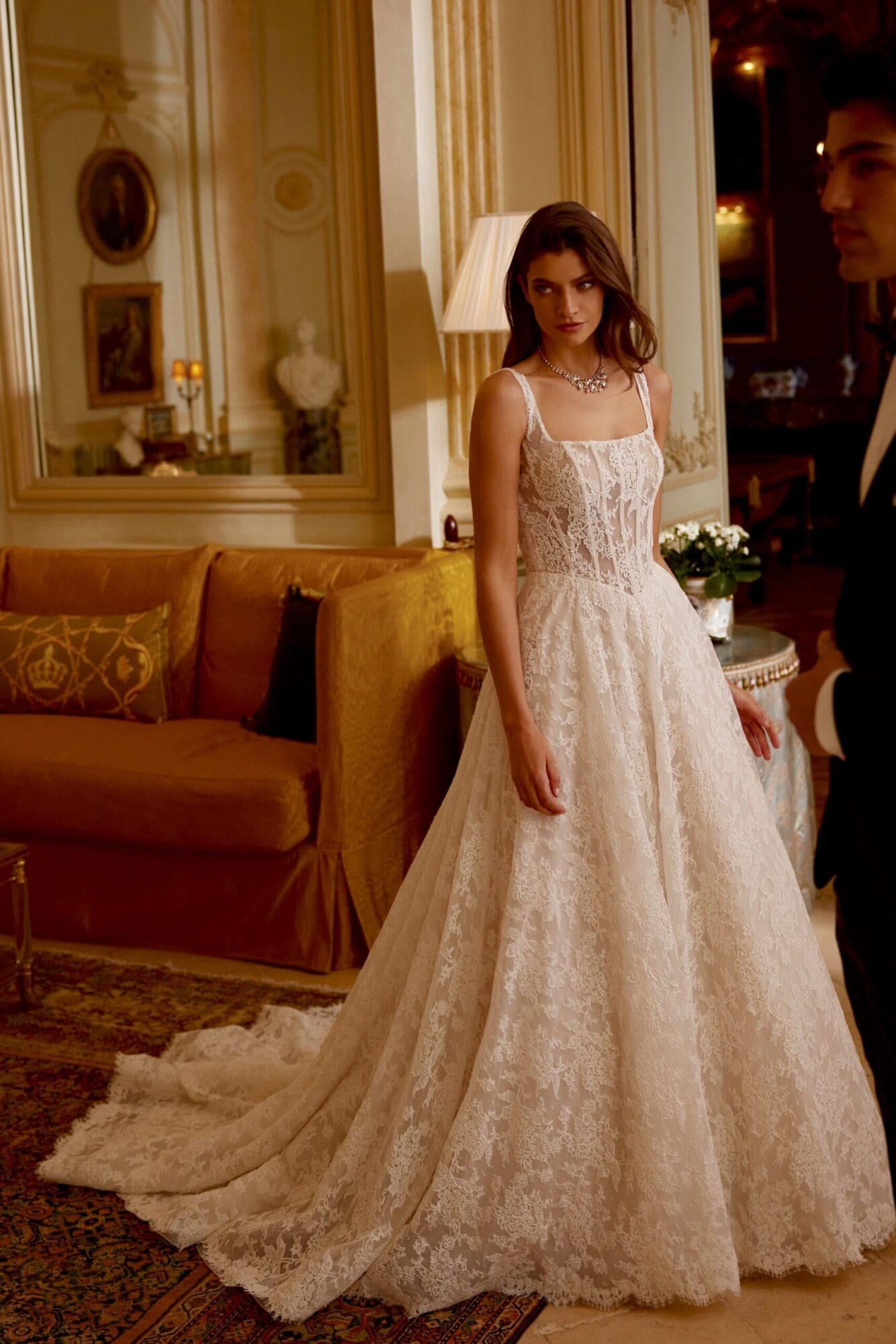1723580858577-88423_Joelle_FF_Justin-Alexander Joelle, a sand, ivory, and latte A-line bridal gown, crafted from allover corded Alencon lace, featuring a sophisticated square neckline, a sheer corset bodice with exposed boning, and a flattering Basque waist. The gown's scalloped lace trim elegantly finishes the hem and trails into a chapel-length train. A woman with long brown hair wears the gown and a sparkling necklace, looking subtly to her left in a lavish, warm-toned room filled with antique furnishings and mirrors, while a man in a black suit is partially visible on the right.