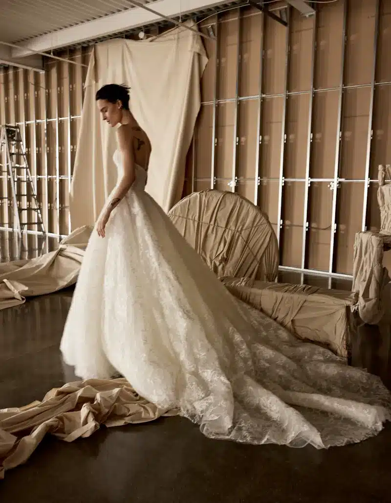 1724957429778-b8b3ffb9-6a92-436c-9fde-e986ba16153a-2 Indigo, an ivory Rivini bridal gown, features a strapless, notched corset bodice and a diaphanous ball gown skirt with a cathedral train, all adorned with intricate beaded 3D floral lace appliqués. A model with short dark hair and tattoos poses elegantly in the gown, looking down, amidst a rustic studio setting with draped beige fabric, exposed wall studs, and cardboard panels.