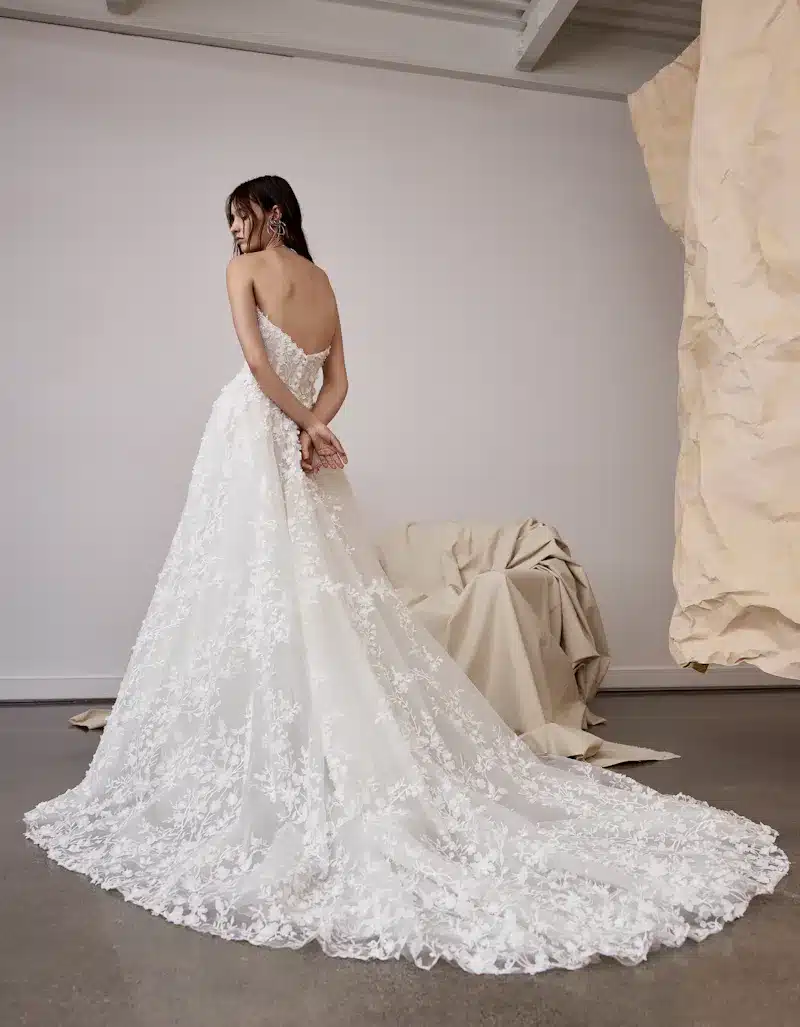 1724957564721-e0ab4335-3978-4893-9a22-cf13773cd240-2 Rhea, an ivory Rivini A-Line bridal gown with a modified sweetheart neckline and Basque waist. This strapless design is crafted from delicate lace and shimmer tulle, featuring floral lace buttons cascading down the back and a long, ethereal train.