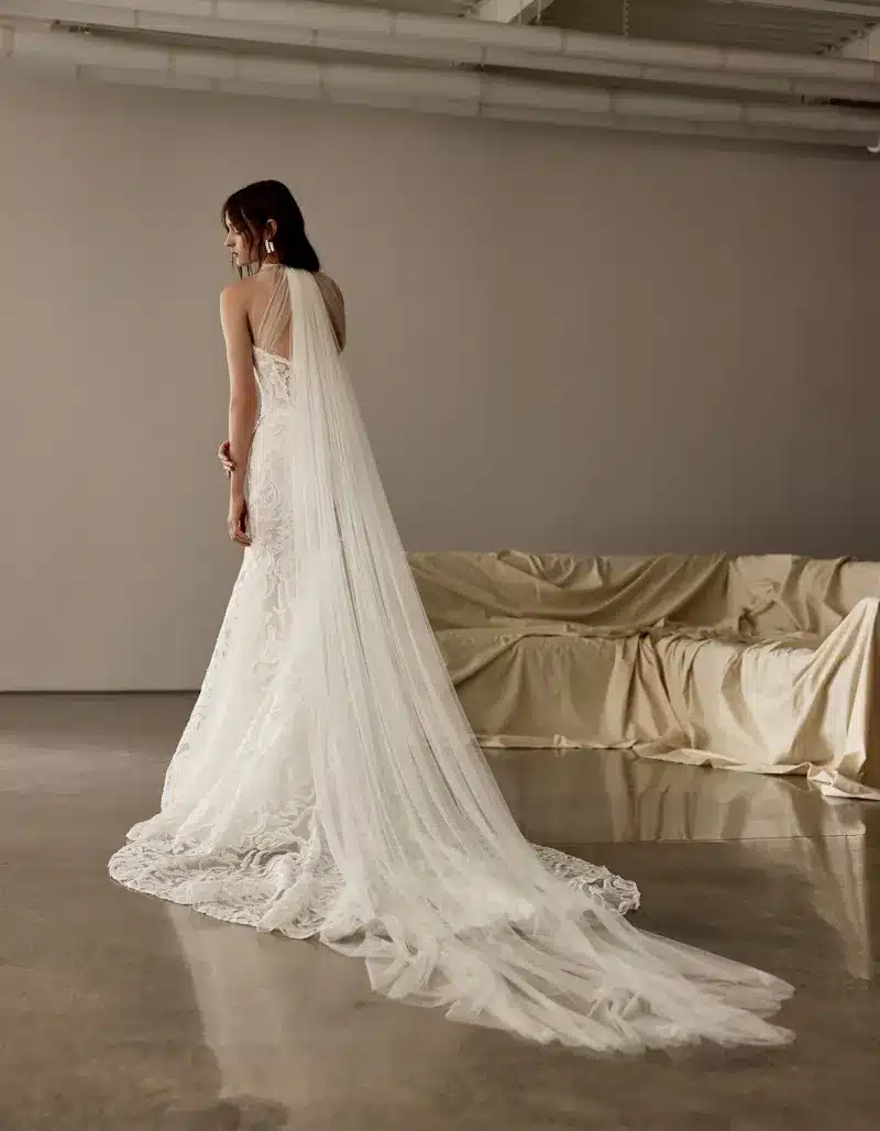 1724957702940-5318235f-bc8e-451a-8f3f-9877d72d3c1e-2 Cameron, an Ivory fit-and-flare bridal gown crafted from beaded embroidered dimensional floral lace. The dress features a structured bodice and a modified sweetheart neckline, with a detachable tulle gilet forming delicate halter-like straps that extend into a stunning watteau train cascading elegantly behind. The model, with dark hair and gold earrings, is captured from behind looking over her left shoulder, standing on a reflective concrete floor in a minimal studio setting with a light brown wall and a draped sofa in the background.