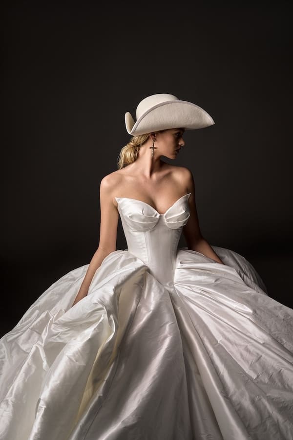 1727278704915-antoinette-gown-side-by-leah-da-gloria-boheme-v-5 Antoinette, a natural white strapless ballgown wedding dress, features a sharply tailored V-shaped corset with sculptural pleats at the bust and a grand, voluminous skirt flowing into a dramatic train. The gown is crafted from crisp silk dupion. A model with blonde hair in a low ponytail, accessorized with a cream cowboy hat and a long cross earring, poses to the right against a dark background.