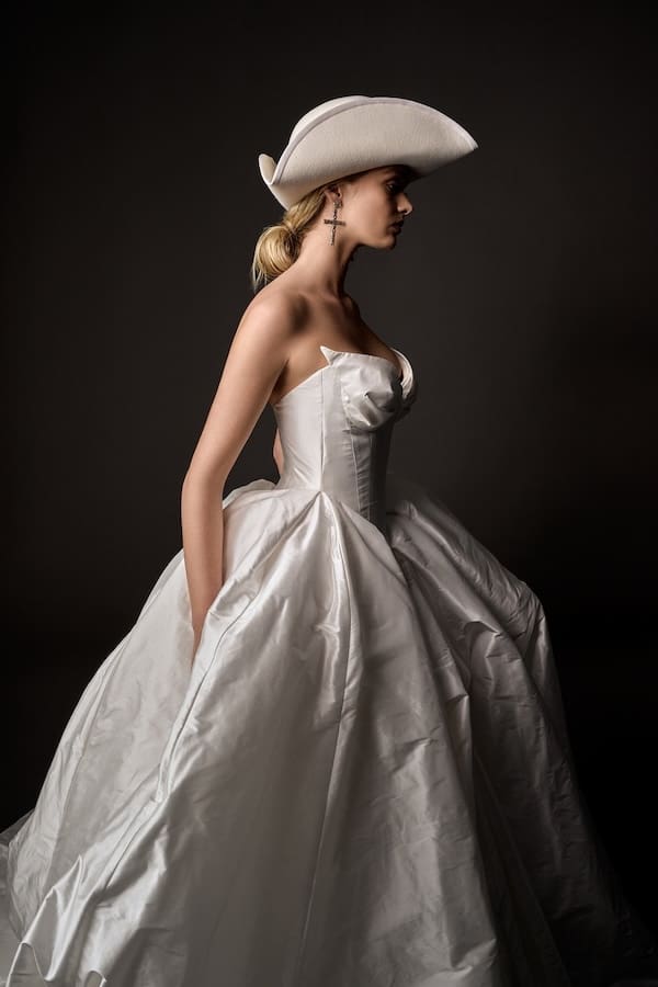 1727278704916-Antoinette-Side-2-Boheme-V-Collection-2 Antoinette, a natural white Leah Da Gloria bridal gown, worn by a model in profile, facing right. The strapless gown features a structured V-shaped corset with sculptural pleats at the bust, a grand, voluminous skirt of crisp silk dupion, and a dramatic train. The model completes the look with a white cowboy hat and cross earrings.
