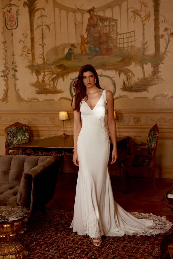 1728399910504-88424-joey-ff-justin-alexander-2 Joey, an ivory and latte stretch crepe fit-and-flare bridal gown by Justin Alexander, featuring a flattering V-neckline, illusion side cutouts and back, and a fully embellished bodice and skirt with sparkling sequin lace appliqués that extend to the chiffon side godets and a chapel-length train. A woman with long dark hair models the dress while standing in an elegant room with a classic wall mural.