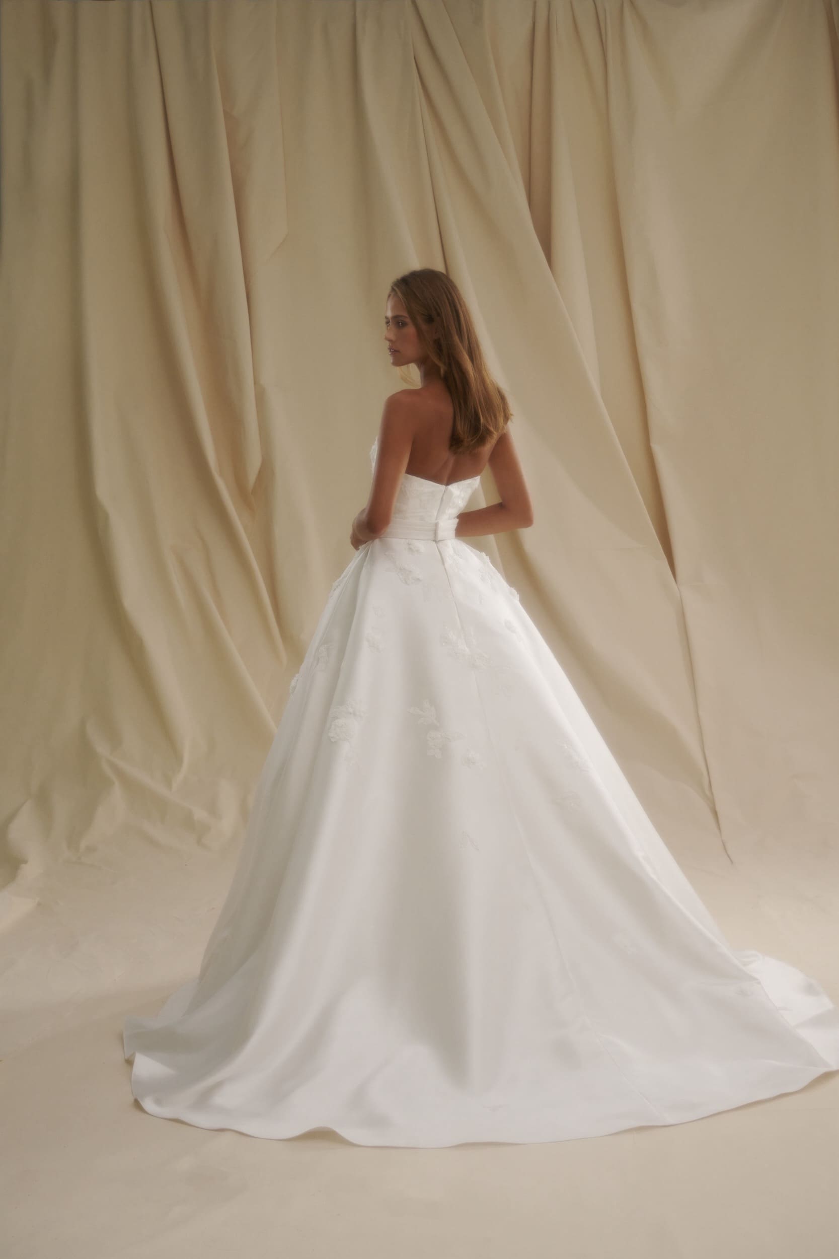 1729715390201-image-1-2 SE027, a Diamond White strapless A-line bridal gown crafted from luxurious mikado fabric, featuring delicate 3D floral appliqués on its voluminous skirt and a cinched waist with a belt. The gown is worn by a model, seen from the back looking over her left shoulder, against a softly draped, neutral-toned fabric background.