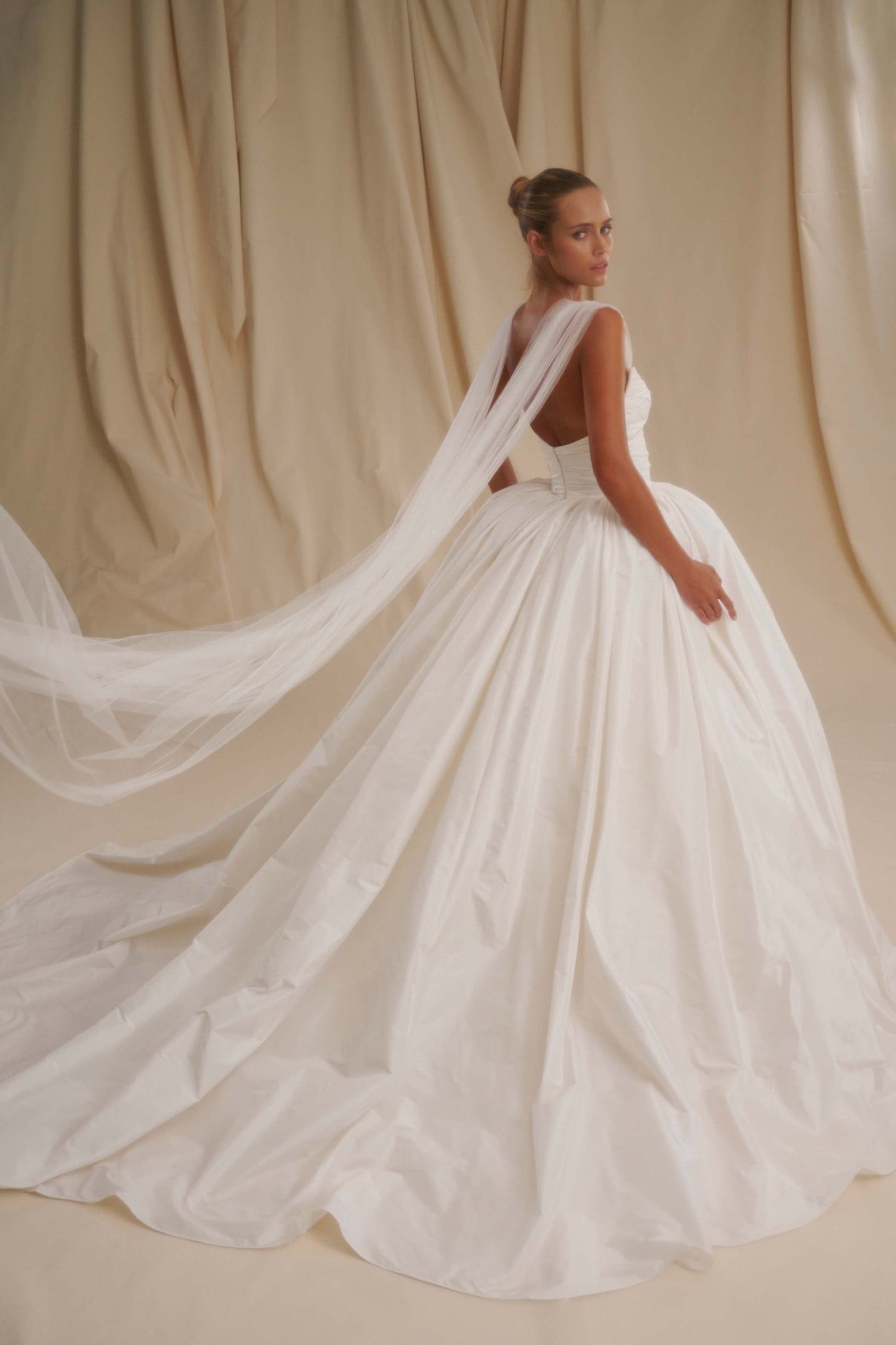 1729716222490-image-3-2 Senstudio, an ivory strapless taffeta bridal ball gown, features a hand-draped sweetheart bodice with ruching and a voluminous skirt. A model, seen from behind and looking over her left shoulder, wears the elegant dress with sheer tulle draped over her shoulders, flowing behind her.
