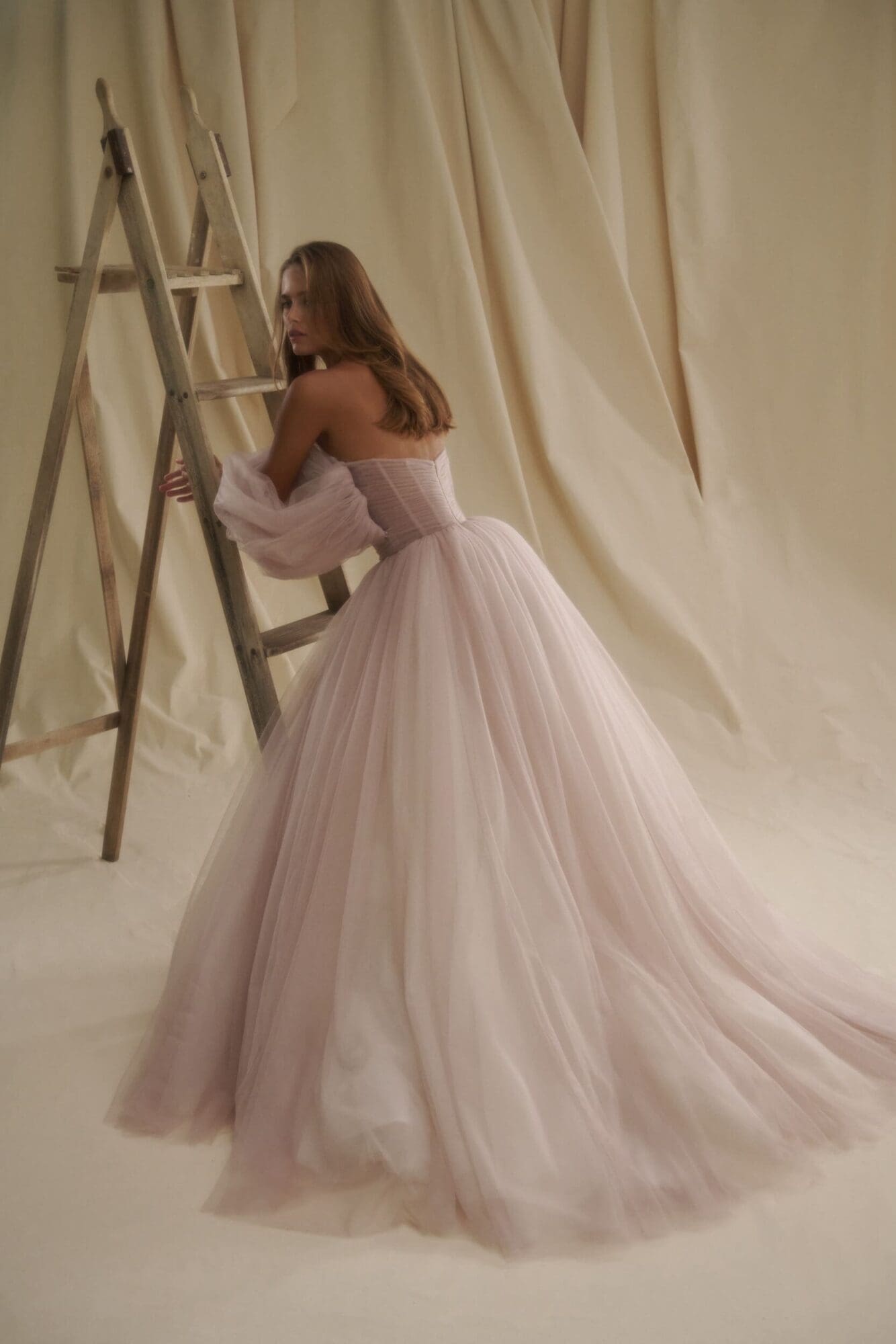 1729716377387-image-4-2 Senstudio's SE008, an ivory (with a soft lavender tint) A-line bridal gown, features a strapless ruched bodice and dramatic billowy tulle sleeves. The chapel-length voluminous skirt flows gracefully as the model poses beside a rustic wooden ladder against a draped fabric backdrop, looking over her shoulder.
