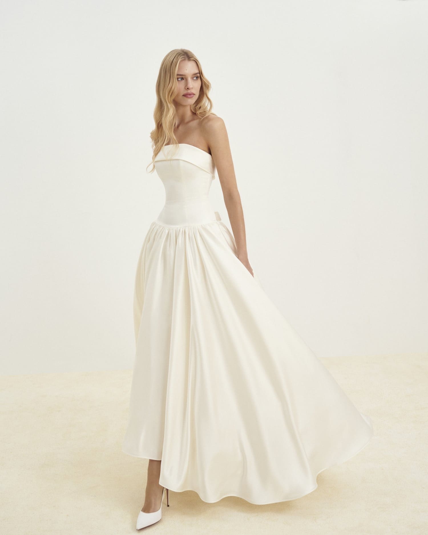 1729782974978-image-5-2 SE038, a cream satin high-low bridal gown by Senstudio. This strapless A-line dress features a folded neckline, a structured corset bodice, and a dropped waist leading to a voluminous skirt. Modeled by a blonde woman wearing white heels, the dress blends modern chic with playful elegance.