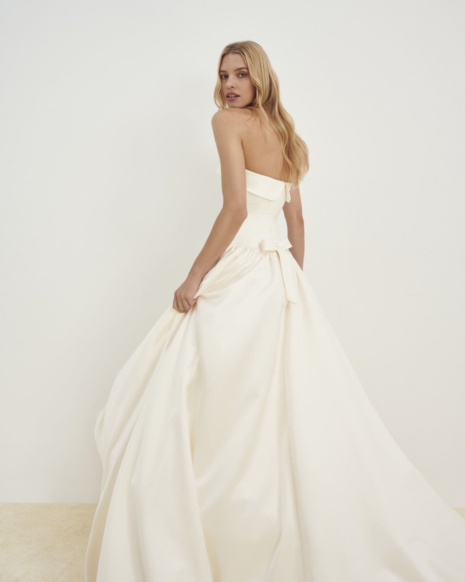 1729782974982-image-6-2 SE038, a cream strapless satin high-low wedding gown by Senstudio, is modeled by a blonde woman looking over her shoulder. The dress features a chic fold-over band at the bodice neckline, a dropped waist, and an A-line skirt with a charming bow detail at the back. She gently holds the flowing, luxurious satin fabric, highlighting its movement against a minimalist white background.