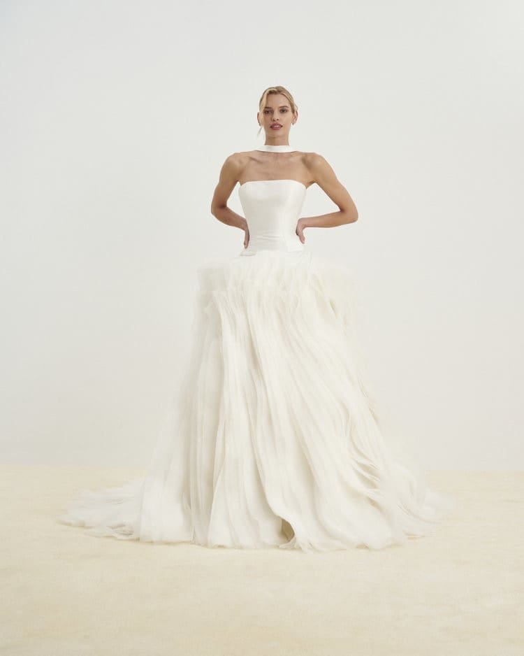 1729783905532-image-11-2 Senstudio Diamond White bridal gown, a strapless design featuring a sleek mikado bodice and a dramatic dropped waist that transitions into a voluminous, sculptural organza skirt. The skirt cascades in ethereal layers, creating soft, flowing shapes that embody modern sophistication and whimsical romance. A woman with blonde hair models the gown, standing with hands on hips against a light background.