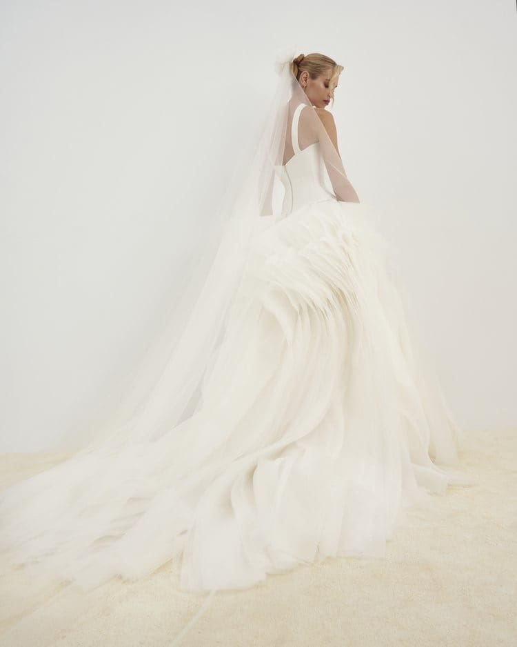1729783905534-image-12-2 SE046, a Diamond White bridal gown by Senstudio, features a sleek mikado bodice with wide straps and a dropped waist, flowing into a dramatically voluminous A-line skirt made of sculptural organza with ethereal, cascading layers and a long train. The bride, seen from behind wearing a veil, looks gracefully over her shoulder, embodying modern sophistication and whimsical romance.