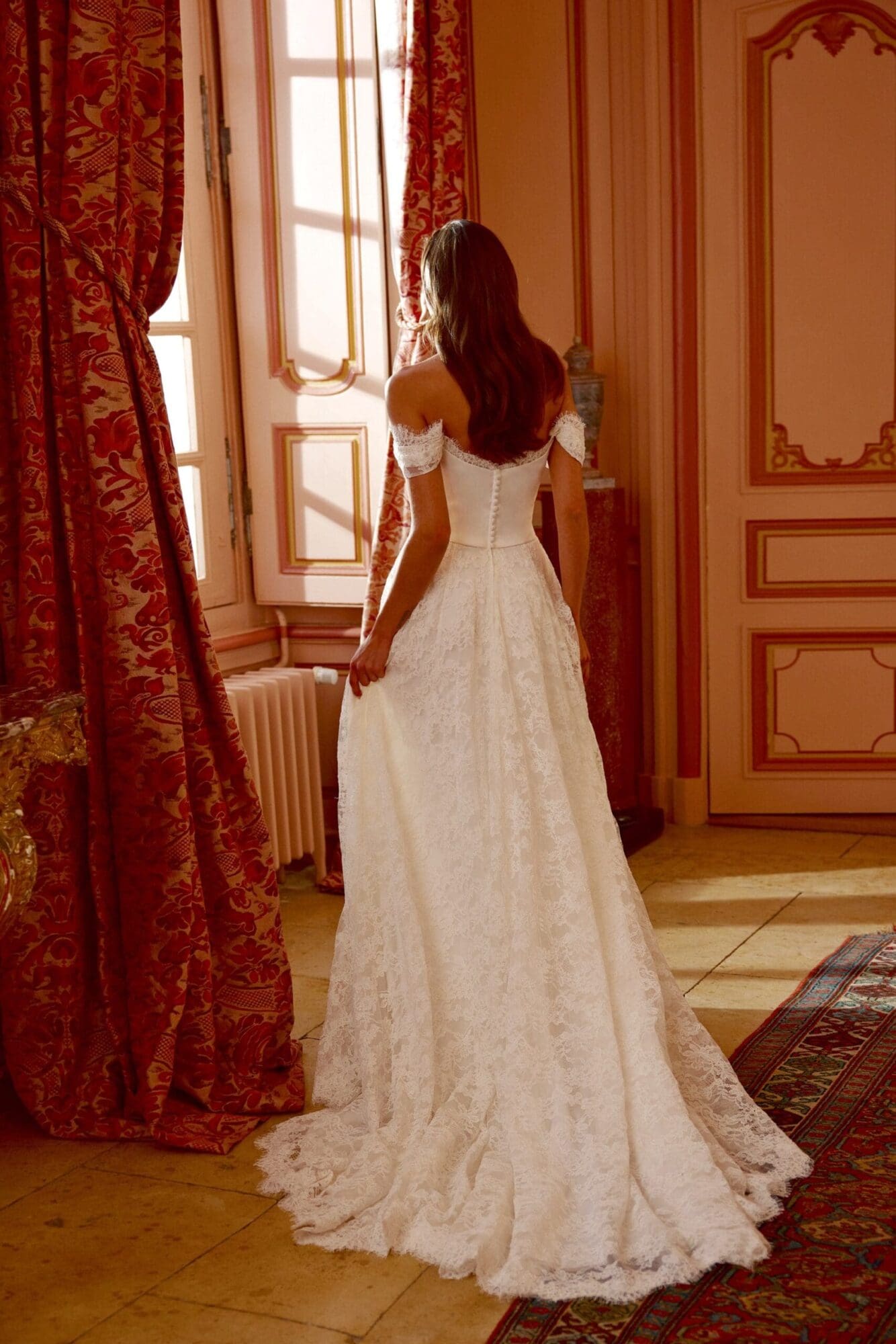 1730305458590-image-16-2 Joyce, an ivory/ivory/latte A-line bridal gown by Justin Alexander, is seen from the back on a bride gazing out a sunlit window in a stately room. The gown features a structured satin corset bodice with a flattering Basque waist and a trail of buttons down the back. It is adorned with exquisite corded Alencon lace, including soft, draped detachable off-the-shoulder straps and an allover lace skirt with a scalloped hemline and a chapel-length train. The room has red damask curtains, ornate wall panels, and a patterned rug.