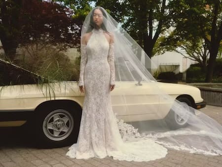 1737657866986-daisy-fw25-alyne-3 Daisy, an ivory and nude Chantilly corded lace halter sheath gown by Alyne, features a high neckline and delicate cold-shoulder lace sleeves. The model stands elegantly beside a vintage cream car, with her long, sheer chapel veil gracefully draped over the vehicle and trailing onto the paved ground in an outdoor setting.