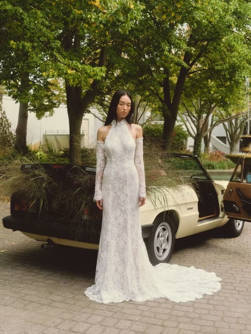 1737658043025-daisy_w25_alyne2-1 Daisy, an ivory and nude Chantilly corded lace halter sheath gown, is modeled by a woman with long dark hair, standing elegantly outdoors. The gown features a high halter neckline, a low back adorned with a handmade organza flower and cascading bow, delicate lace sleeves, and covered buttons extending down its chapel train. Behind her is a vintage cream convertible car, its back filled with dried grasses, set against a backdrop of lush green trees and a fountain.