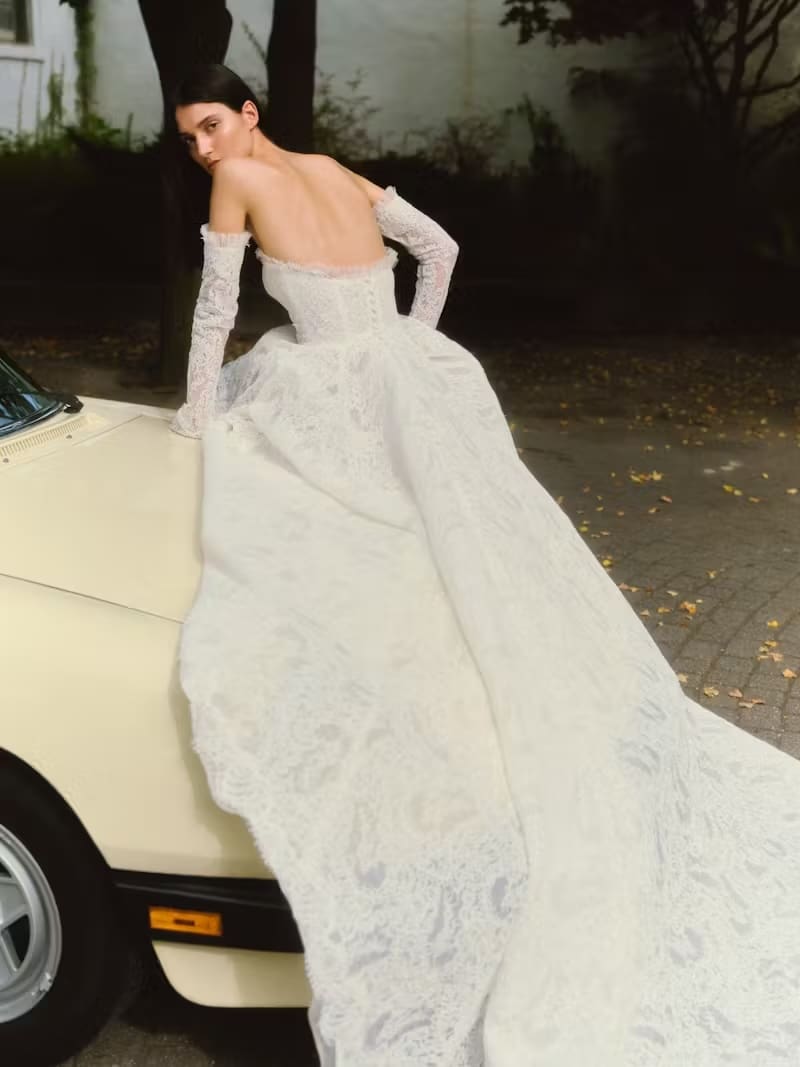 1737659138714-magnolia-alyne3-4 Magnolia, an ivory Alyne strapless grand ballgown crafted from corded lace with a dramatic silhouette and a long train, worn by a model with elegant detachable lace sleeves. The model is gracefully posed, leaning on the hood of a vintage light-colored car in a dimly lit outdoor setting, looking over her shoulder.