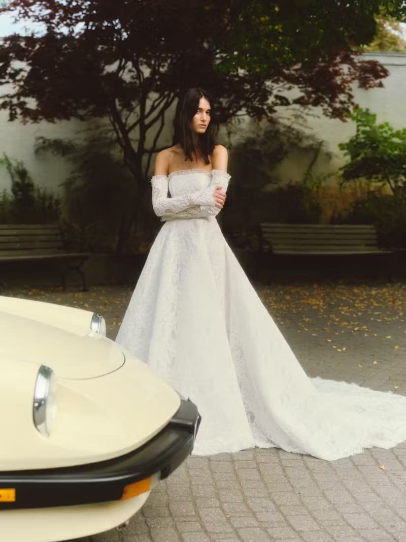 1737659138714-magnolia_alyne2 Magnolia, an ivory strapless ballgown crafted from exquisite corded lace and micro-pleated tulle, features a dramatic silhouette, detachable lace sleeves, and a row of buttons down the back. A model with dark hair wears the gown while standing next to a vintage cream-colored car in an outdoor setting with trees and benches.