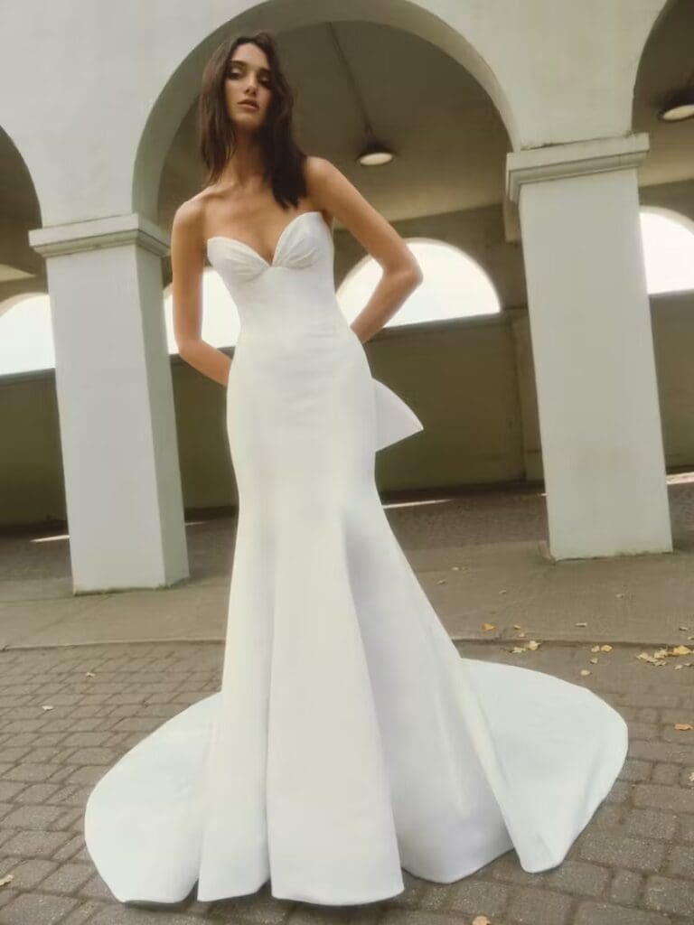 Jasmine, an ivory soft satin fit-and-flare bridal gown with a cathedral train, featuring a strapless draped cat-eye sweetheart neckline, a structured bodice, and a flattering Basque waist. The gown is detailed with a sheer back adorned with boning and delicate buttons trailing to the hem, completed by a dramatic double bow. The model poses elegantly under white archways.