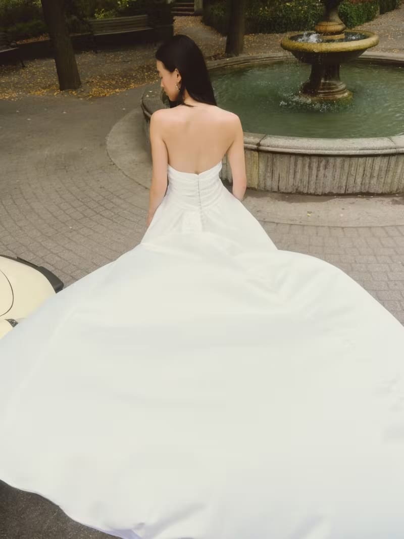 1737661288367-alyssa-alyne3-2 Alyssa, an off-white strapless dropped-waist ballgown by Alyne, featuring a draped bodice, a voluminous pleated skirt with hidden pockets, and a cathedral-length train with a row of delicate buttons cascading down the back to the hem. The gown is worn by a bride, viewed from behind, looking towards a fountain in a park setting, with a part of a vintage car visible on the left.