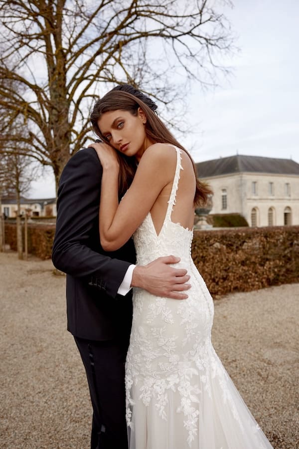 1738944238311-jazmin-ja1-1 Jazmin, a Sand/Ivory/Latte trumpet wedding gown by Justin Alexander, showcasing a chic square neckline, spaghetti straps, and a fitted silhouette adorned with intricate beading, sequins, and lace appliques that flow into a chapel-length train. The bride, with long dark hair, is embraced by a person in a black suit, gazing towards the viewer in an outdoor setting featuring a historic building.