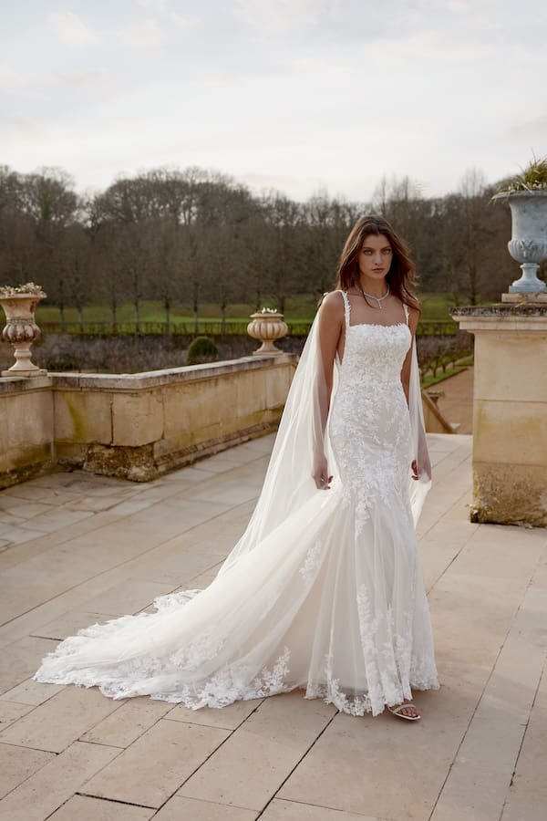 1738944238313-jazmin-ja3-1 Jazmin, a Sand/Ivory/Latte modern and romantic trumpet bridal gown, featuring a chic square neckline and delicate spaghetti straps. The fitted silhouette is adorned with intricate beading and sequins over lace, flowing into a shimmering chapel-length train. A model wears the gown on a grand outdoor stone terrace with balustrades and bare trees in the background.
