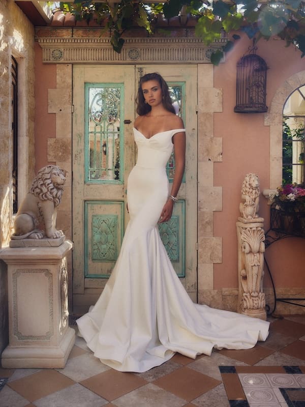 1738944952185-Elora_LDG1-2 Elora, a white crepe bridal gown by Leah Da Gloria, is modeled by a woman with long brown hair, standing in a rustic, sunlit Mediterranean courtyard. The dress features a draped off-the-shoulder neckline, a form-skimming silhouette, and a dramatic mermaid skirt with a train. Behind her is an ornate pale blue wooden door, flanked by carved stone lion statues.