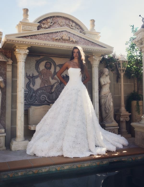 1738945565552-bijou-ldg3-4 Bijou, a white strapless ball gown wedding dress with a dropped waist and intricate 3D floral appliqués adorning its voluminous skirt and long cathedral train. The dress is modeled outdoors by a woman with long brown hair, standing in front of an ornate classical building featuring columns, archways, and a mosaic depicting a figure on a chariot. To her right, a classical statue stands beside a decorative lamp post. The scene is bright and grand, with a pool in the foreground. Designed by Leah Da Gloria.