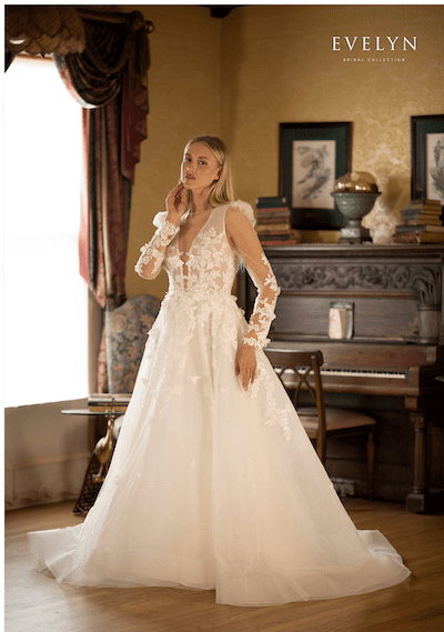 1738952968574-jolene-evelynbridal Delia, an Ivory/Champagne Ball Gown wedding dress with a flattering plunge V-neckline, illusion long sleeves adorned with lace and sparkle, and a voluminous tulle skirt featuring soft flat lace appliqués, shimmering sequins, and delicate 3D embroidered flowers. The gown has an open back with a button closure and a graceful cathedral train, modeled by a blonde woman in a stately room.