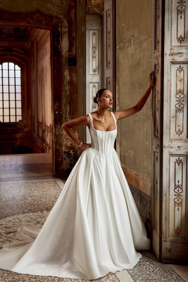 1740168162466-88440-keegan-ff-justin-alexander-1 Keegan, an ivory stretch mikado ball gown, features a square neckline and a structured corset bodice with exposed boning and a flattering Basque waistline. Embroidered sequined lace adorns the neckline, waistline, bodice sides, and straps, continuing onto the sides of the skirt and down the center back. The voluminous skirt includes pockets. A soft V-back is completed with buttons trailing down the cut-out cathedral train, accented with more sequined lace. A Black model with her hair pulled back stands in an ornate, old-world hallway, leaning against a decorative wooden door with one arm raised, looking off to the side.