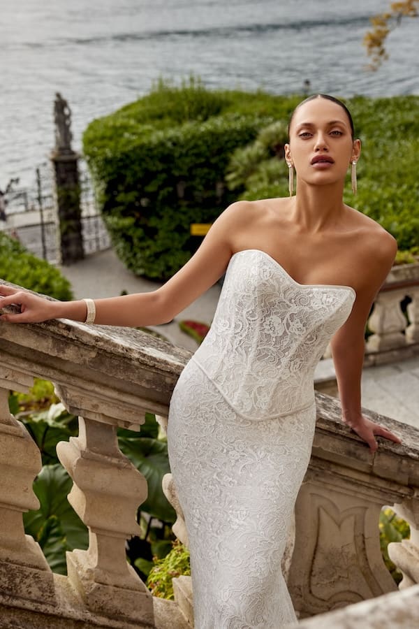 1740168669442-88457bod_keeley-bodice_fc_justin-alexander_1 Keeley, a two-piece bridal gown in sand, ivory, and latte, featuring a corded lace corset bodice with a modified sweetheart neckline, satin piping along the basque waist, and exposed boning. It is paired with a matching fit-and-flare corded lace skirt. The bride poses elegantly, leaning on a classical stone balustrade with a lush green garden and a tranquil body of water in the background.