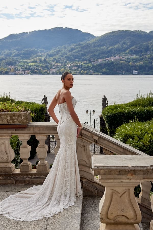 1740168674556-88457bod-keeley-bodice-fb-justin-alexander-2 Keeley, a Sand/Ivory/Latte 2-piece bridal gown by Justin Alexander, features a strapless, modified sweetheart corset bodice with demi-sheer exposed boning and a basque waist, paired with an allover corded lace fit-and-flare skirt that flows into a chapel-length train with buttons and eyelash lace trim. The bride stands elegantly on a grand stone balcony, looking over her shoulder at the viewer, with a serene lake bordered by mountains and distant towns in the background.