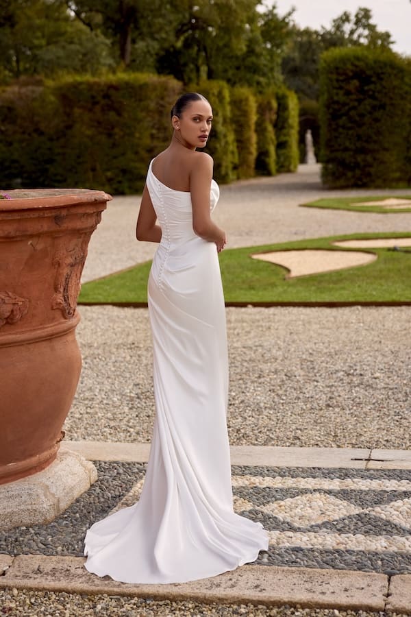 1740171614959-88445-knox-fb-justin-alexander Knox, an ivory charmeuse fit-and-flare gown, featuring a draped one-shoulder neckline, a structured corset bodice with a basque waist, and elegant buttons extending down the back, is worn by a model with her hair pulled back, looking over her shoulder at the viewer, standing in a formal garden with gravel paths, manicured hedges, and large terracotta planters.