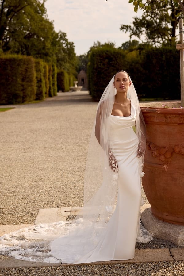 1740171614962-88445-TG1086L120_Knox_FF_Justin-Alexander_1-1 Knox, an ivory charmeuse fit-and-flare wedding gown by Justin Alexander, featuring a draped cowl neckline and elegant draping across the bodice and skirt. The gown flows into a chapel-length train, adorned with subtle floral appliqués that match the long, sheer veil worn by the bride. She stands posed on a gravel path in a formal garden, alongside a large terracotta pot, with sculpted hedges and a distant fountain adding to the serene backdrop.