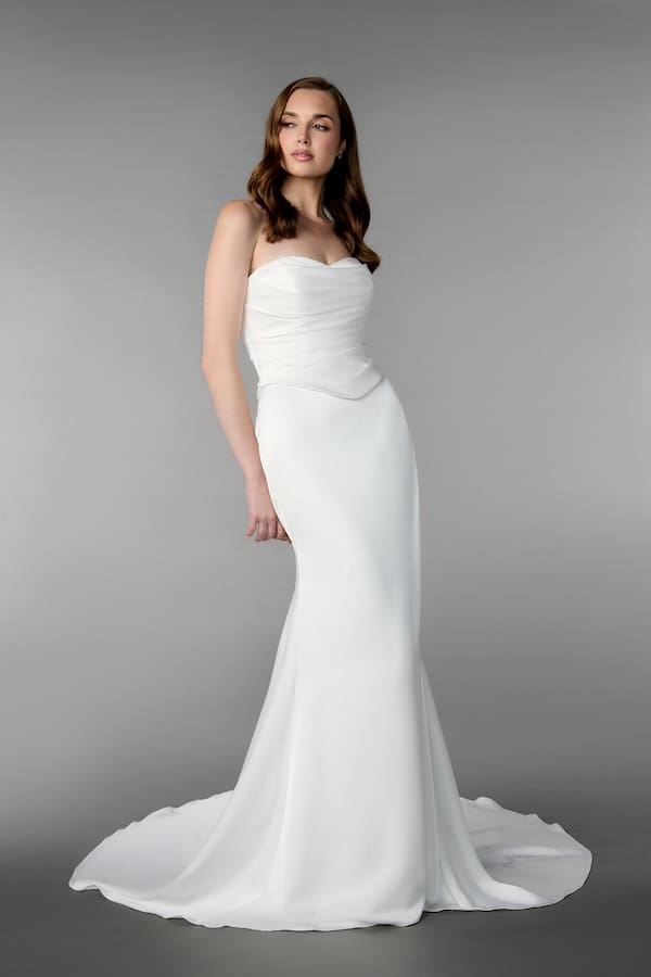 1740172944729-RNG_55233 Aphrodite, an ivory charmeuse fit-and-flare bridal gown, modeled by a woman against a grey background. The gown features a sweetheart neckline, a beautifully draped and ruched bodice, and a sleek skirt flowing into a chapel-length train. Designed by Adorn Nashville, it includes a lace-up corset back.