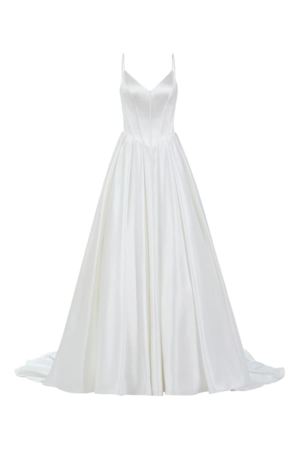 1740173332639-RNG_55234LND Lucy, an ivory satin A-line bridal gown with a sleek V-neckline and delicate spaghetti straps. The bodice features corset boning and a classic basque waistline, leading to a voluminous pleated skirt with hidden pockets. The gown is finished with a V-back and a row of buttons cascading to a chapel-length train. Designed by Adorn Nashville.