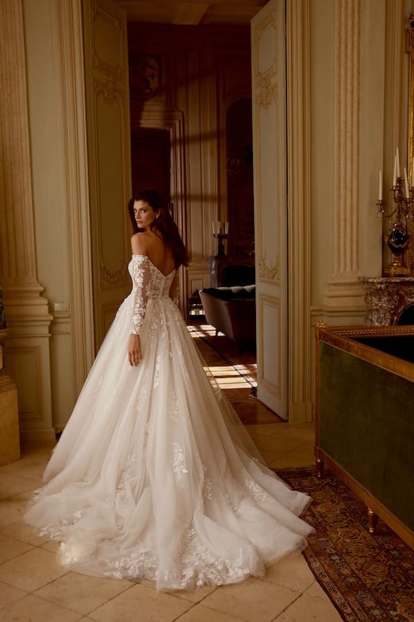 1743018273248-JAB-Janelle-FB-1 Janelle, a Sand/Ivory/Latte glitter tulle ball gown by Justin Alexander. This elegant bridal gown features a straight off-the-shoulder neckline and long sheer sleeves, adorned with delicate sequin lace appliqués that cascade down the bodice and sleeves onto the shimmering tulle skirt. The sheer bodice, with its exposed boning and draped tulle, leads to a gentle V-back detailed with a row of buttons. The voluminous skirt flows into a chapel-length train. A bride models the dress from the back, looking over her shoulder, in an opulent room with warm lighting.