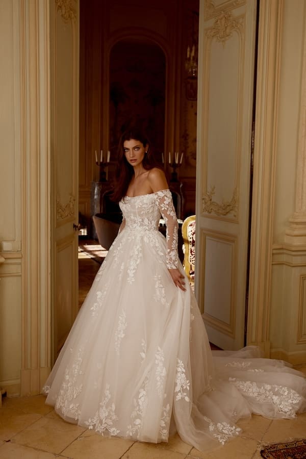 1743018273249-JAB_Janelle-2 Janelle, a Sand/Ivory/Latte glitter tulle ball gown featuring a straight off-the-shoulder neckline and long sheer sleeves. Delicate sequin lace appliqués adorn the sheer bodice with exposed boning, cascading onto the voluminous tulle skirt and chapel-length train. A model in the gown stands elegantly in an ornate room with open double doors.